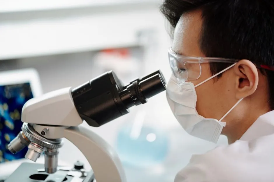 A researcher in a face mask and safety glasses looking through a microscope