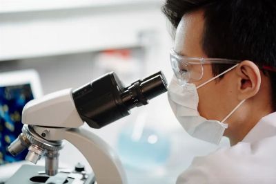 Scientist wearing face mask and goggles examining samples through a microscope in a lab.