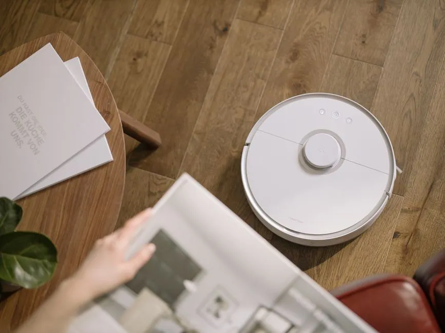 A round white robot vacuum on a wooden floor beside a low table, with a person's hand reaching towards a magazine.