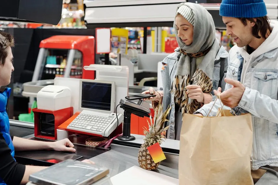 A cashier scans groceries at a supermarket checkout while two shoppers pack a paper bag.