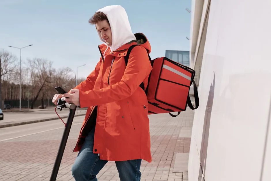 A delivery courier in an orange coat with an unbranded red thermal backpack checking a phone beside a scooter