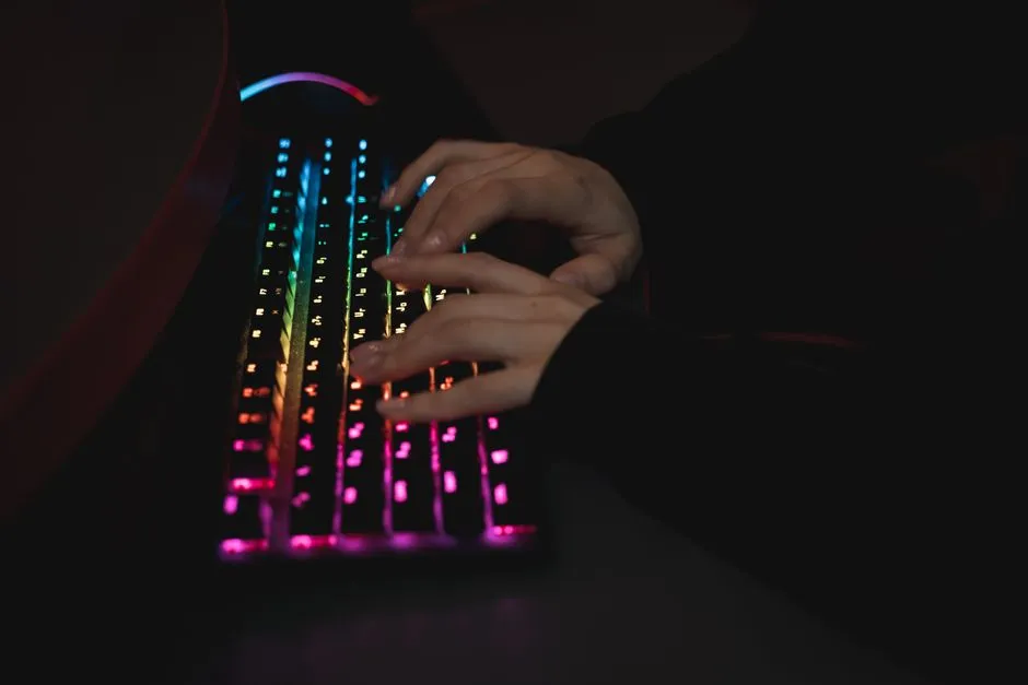 Hands resting on a backlit keyboard in a dark room, the keys glowing in colour
