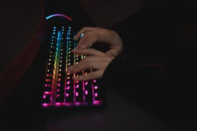 Close-up of hands typing on a colorful RGB backlit mechanical keyboard in a dimly lit environment.