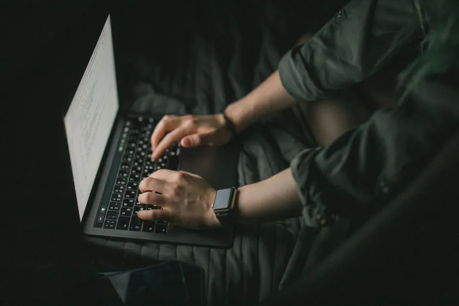 A person sitting cross-legged in low light, typing on a laptop with lines of code visible on the screen