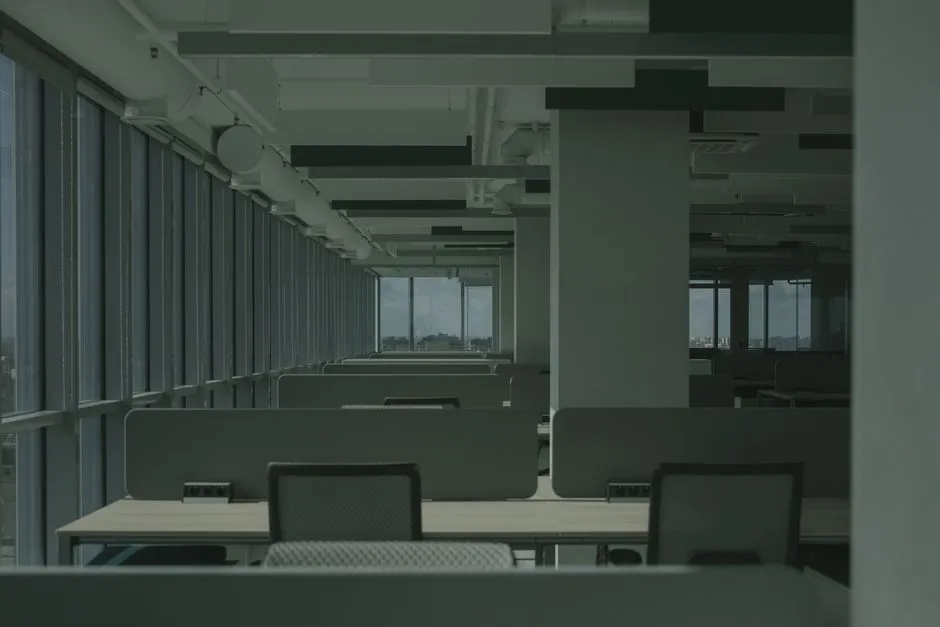 Rows of empty desks and chairs in a dim open-plan office, with daylight coming through a wall of windows