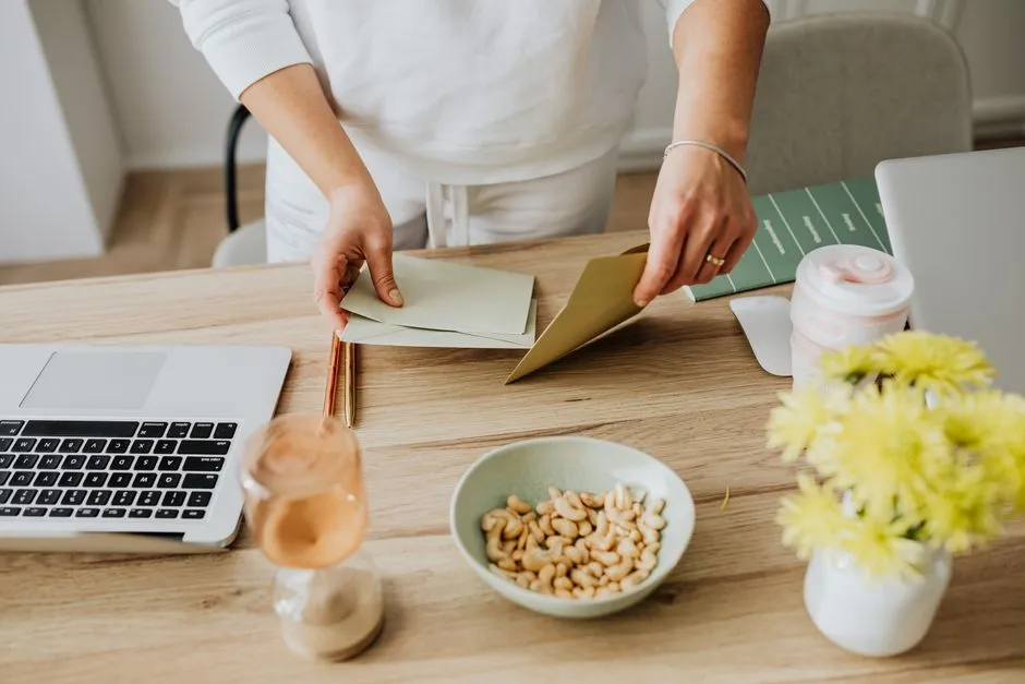 A person at a desk opening a paper document beside a laptop, with a cup and flowers on the table.