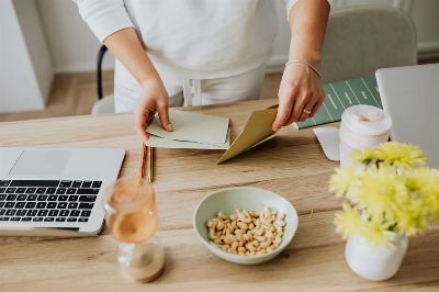 A woman arranges envelopes on a desk with a laptop, cashews, and flowers.