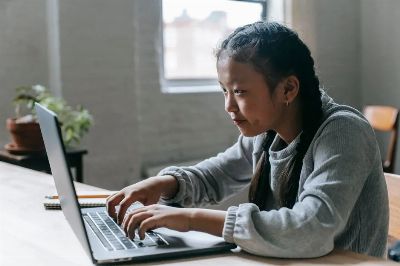 Side view of concentrated Asian child with pigtails sitting at wooden table with textbook and pencil while typing on laptop…