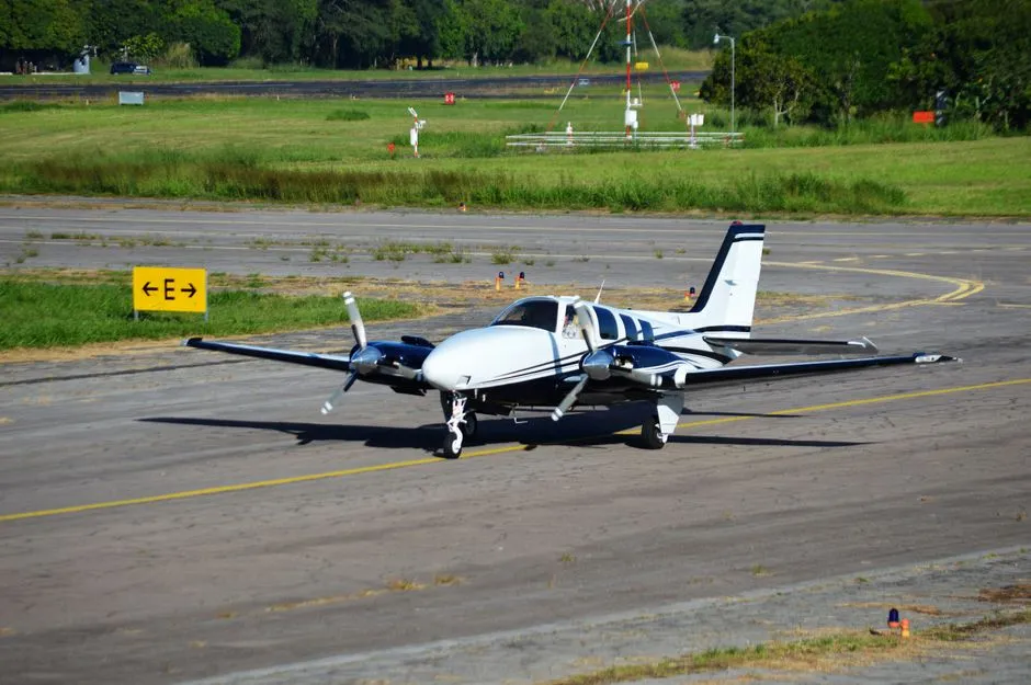 A twin-engine propeller aircraft taxiing across a sunlit runway beside open grass
