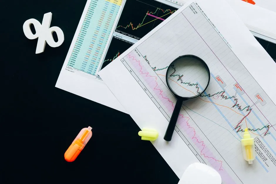 Printed financial charts spread on a dark desk with a magnifying glass held over one line and a percent symbol beside it