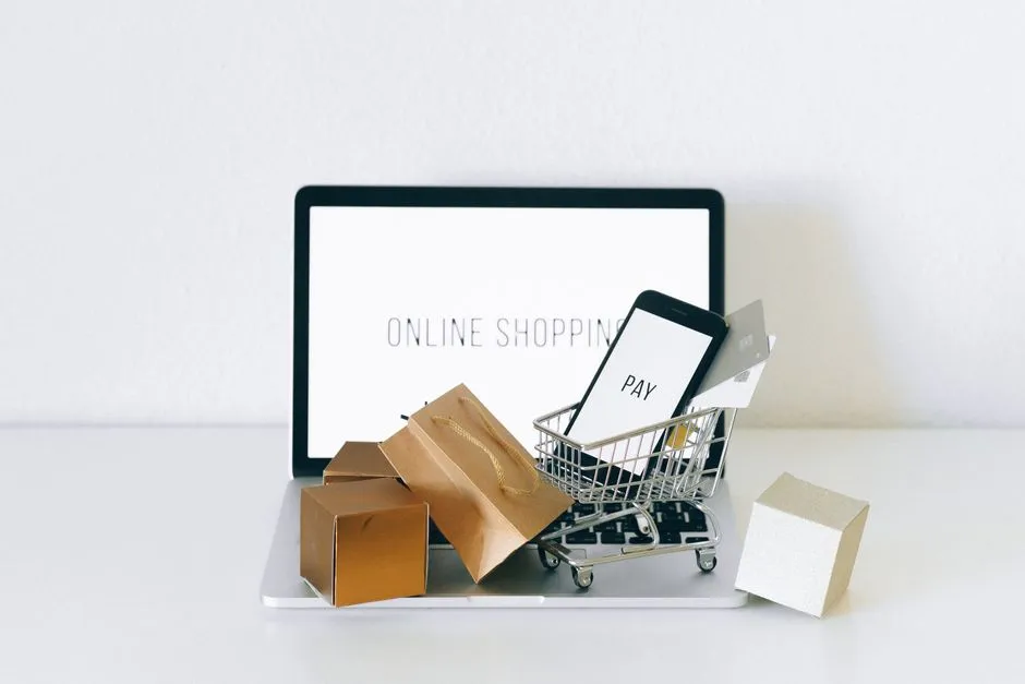A laptop displaying an online shopping page beside a miniature wire trolley holding small parcels.
