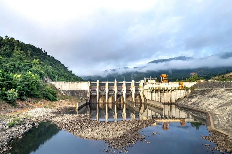 A concrete hydroelectric dam with spillway gates, set in a green river valley under low cloud.