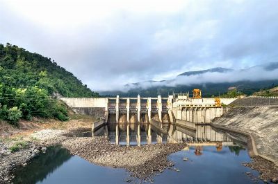 Industrial hydroelectric power plant located near river and stony coast against green mountain range covered with mist on su…