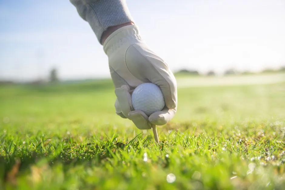 A gloved hand setting a golf ball on a tee in short grass