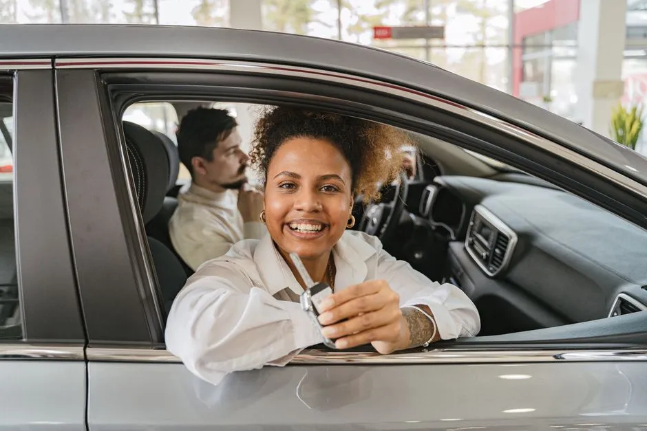 A smiling woman leaning out of a car window at a dealership, holding a set of keys, with a man in the passenger seat