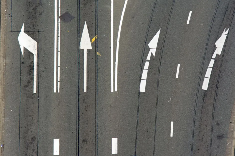 Aerial view of an asphalt road surface painted with large white arrows across five lanes, pointing left, straight ahead and right.