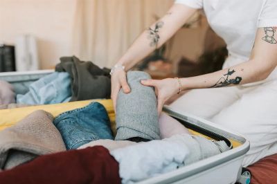 Close-up of a person packing clothes into a suitcase, showing arm tattoos, in a cozy indoor setting.