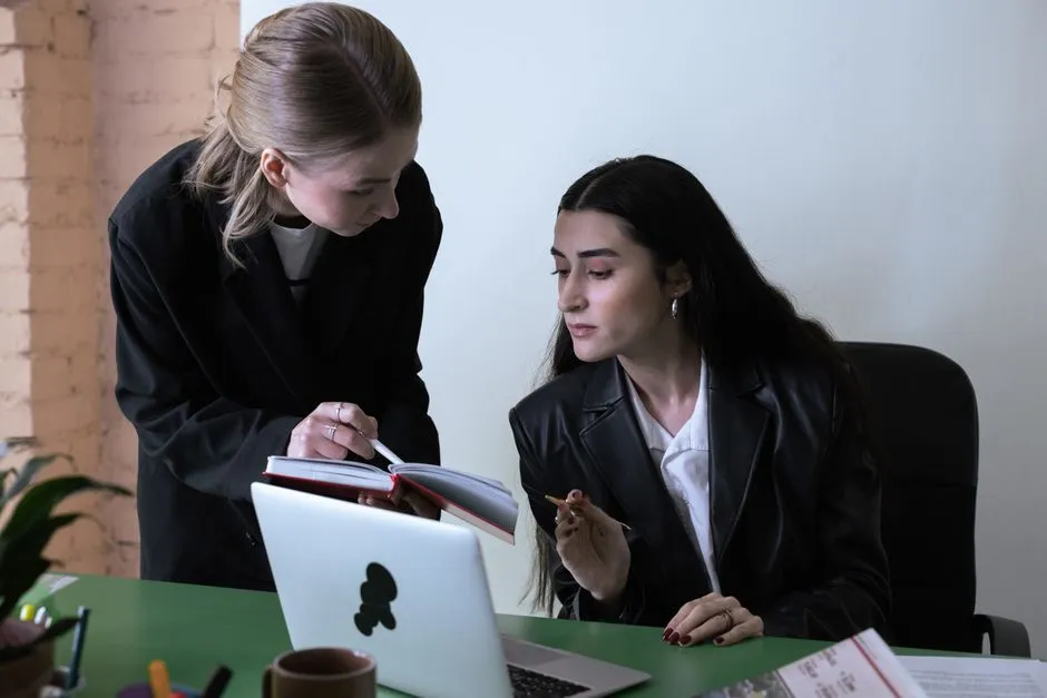 Two colleagues in dark jackets at an office desk, one standing and pointing into an open notebook while the other, seated at a laptop, looks at the same page.