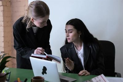 Two female colleagues collaborating at an office desk with a laptop and notebook.