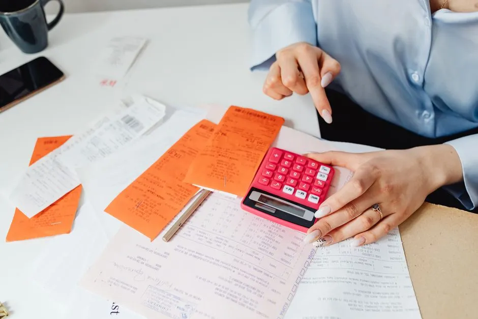 A person at a white desk resting one hand on a pink calculator, with printed slips, receipts and a filled-in form spread out in front of them.