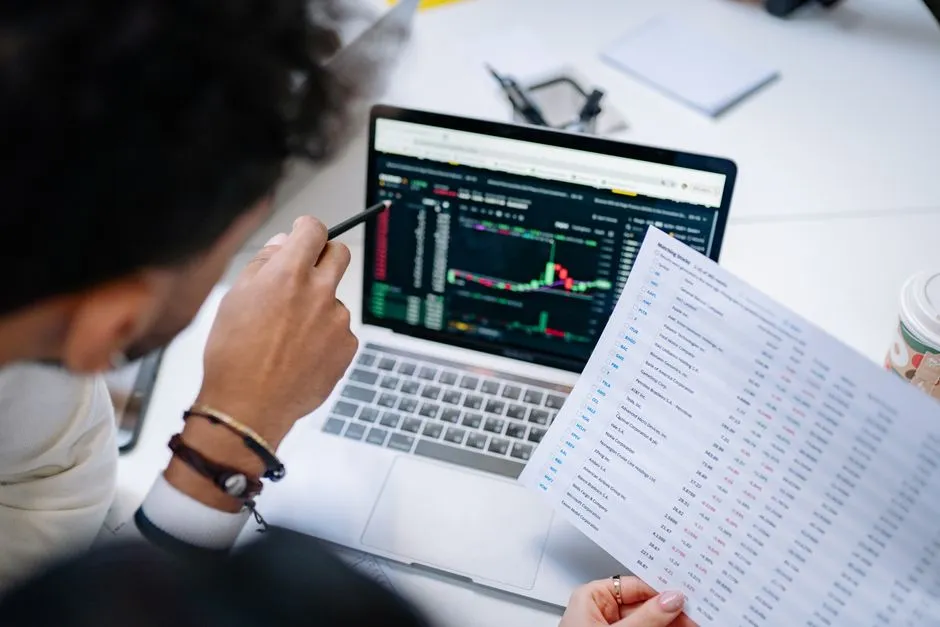 A person at a desk holding a printed sheet of numeric columns in one hand while a laptop beside them shows the same kind of data as a coloured chart.