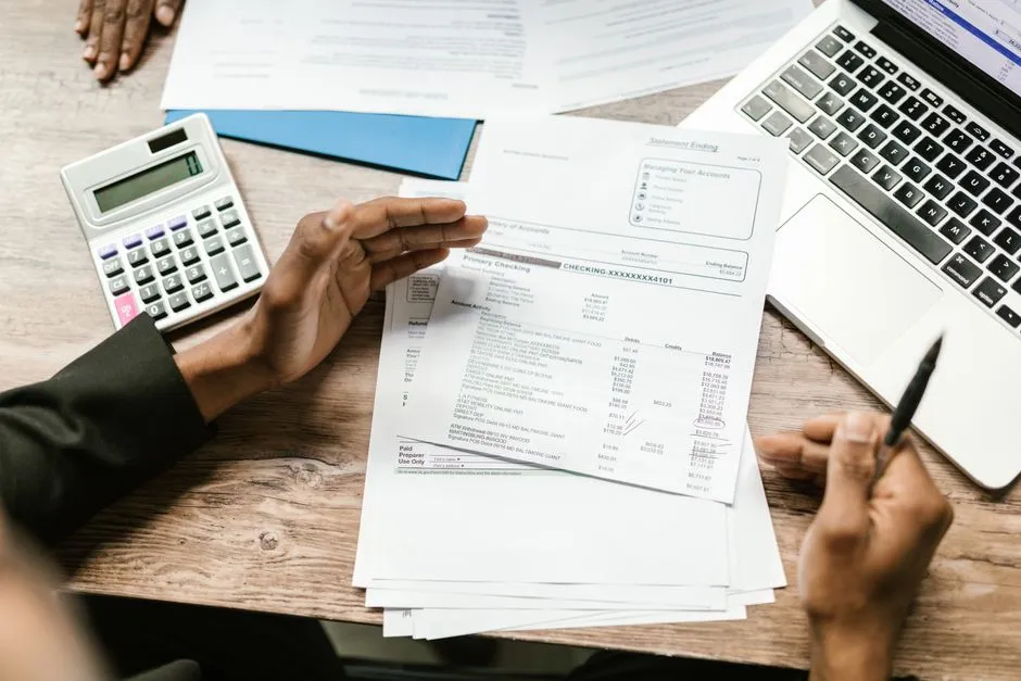 Overhead view of hands examining printed financial statements at a desk, with a calculator, a laptop and a pen