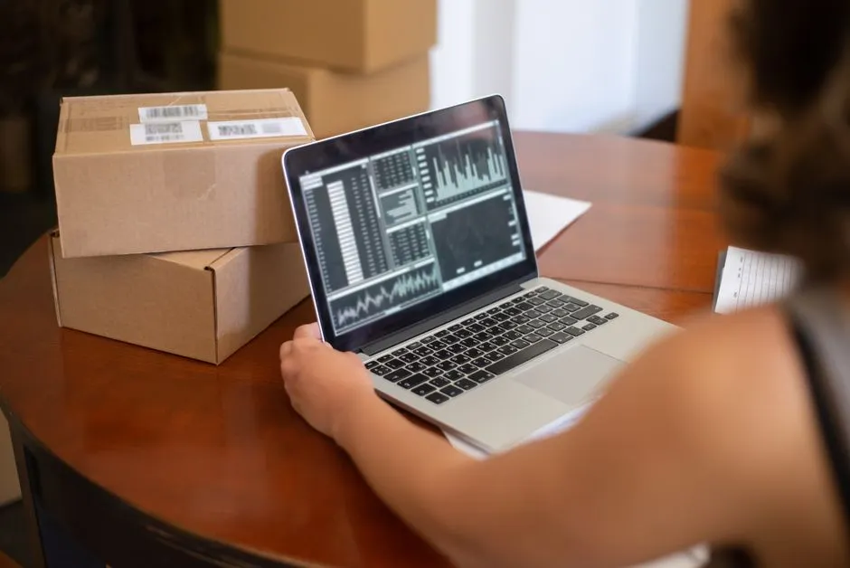 A small-business seller works at a laptop showing performance charts, with stacked cardboard parcels beside it on a wooden table