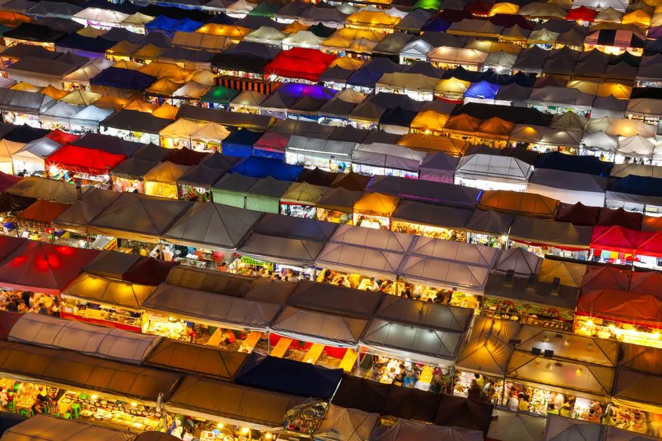 An aerial view of a night market after dark: dozens of rows of stall canopies in red, blue, white and orange, each lit from beneath, packed edge to edge so the individual stalls blur into a grid.