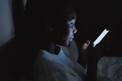 A woman using a smartphone in a dark bedroom, lit by the screen glow at night.