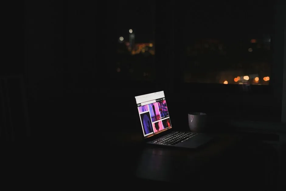 A laptop screen glowing alone in a dark room, with city lights blurred beyond the window