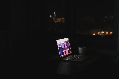 A laptop glows in a dark room at night with a cityscape through the window. Ideal for tech and solitude themes.