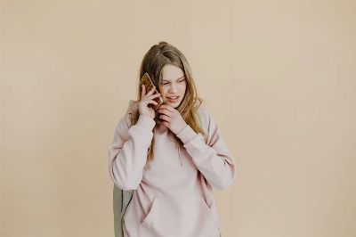 Casual young woman in pink hoodie talking on the phone against a beige background.