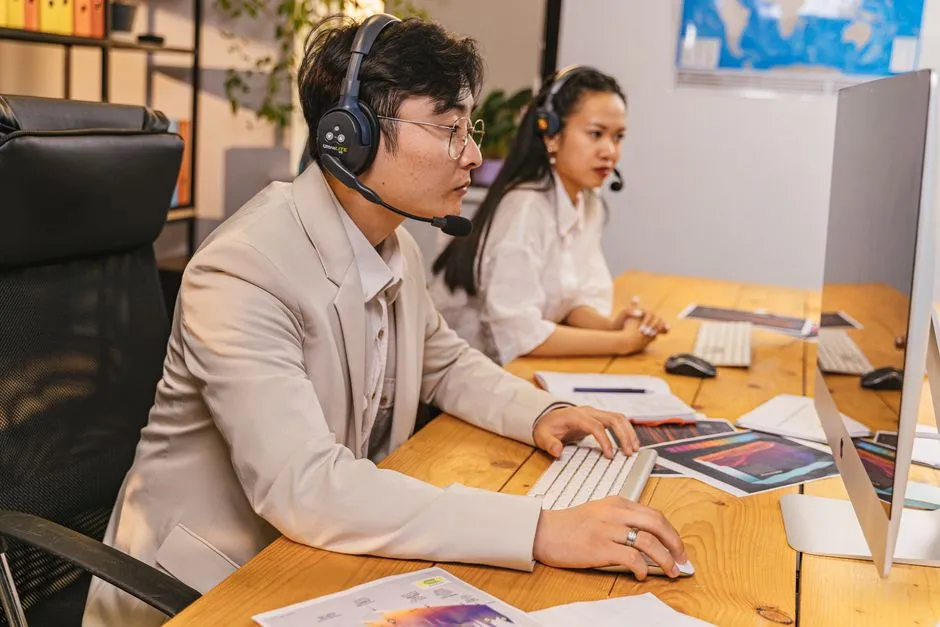 Two call-centre workers in headsets at a shared wooden desk, one typing at a monitor