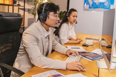Two office workers using computers and headsets in a modern workspace, collaborating on tasks.