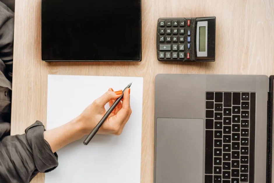 Overhead view of a wooden desk with a laptop, a desk calculator and a closed notebook, and a hand holding a pen over a blank sheet of paper.