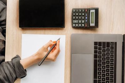 A person writing on paper surrounded by a laptop, tablet, and calculator on an office desk.