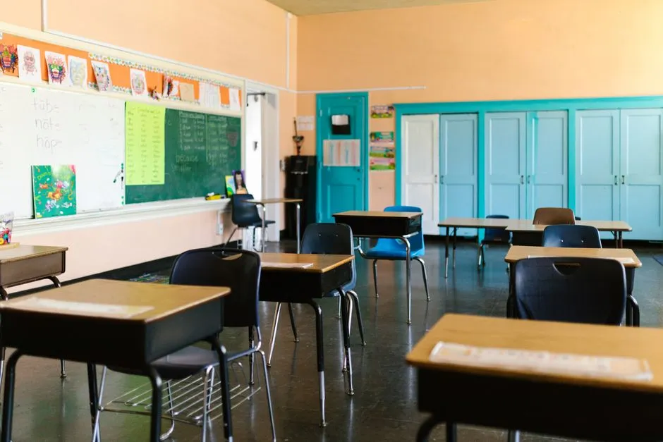 An empty school classroom with rows of wooden desks, a green chalkboard and a line of teal storage cupboards.