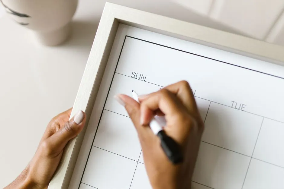 A hand marking a date on a blank weekly wall planner with a pen.
