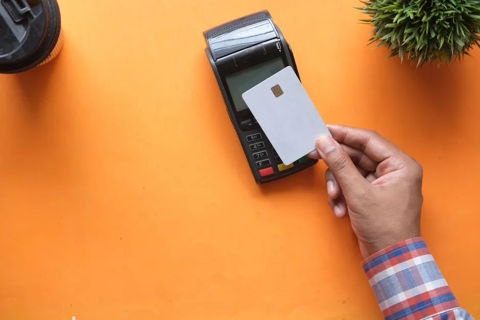 A hand holding a blank payment card above a card terminal on an orange surface