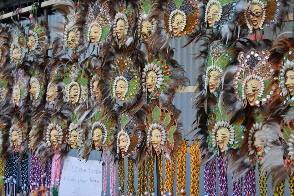 A market stall wall hung with dozens of near-identical festival masks, each a smiling face fringed with feathers and coloured beadwork, repeating in rows so that individual masks are hard to tell apart.