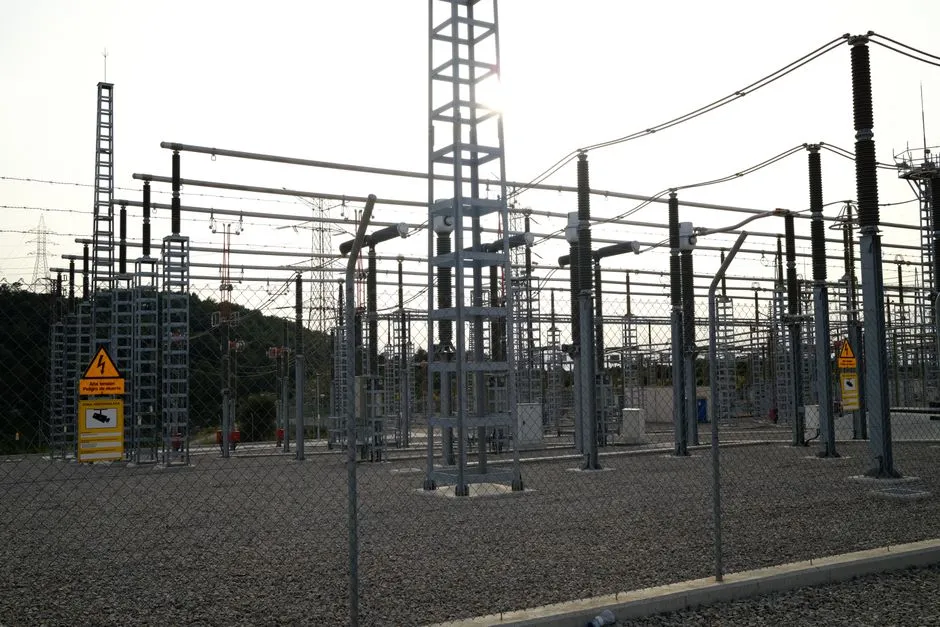 An electrical substation seen through a chain-link fence at dusk, steel gantries and high-voltage insulators backlit by low sun