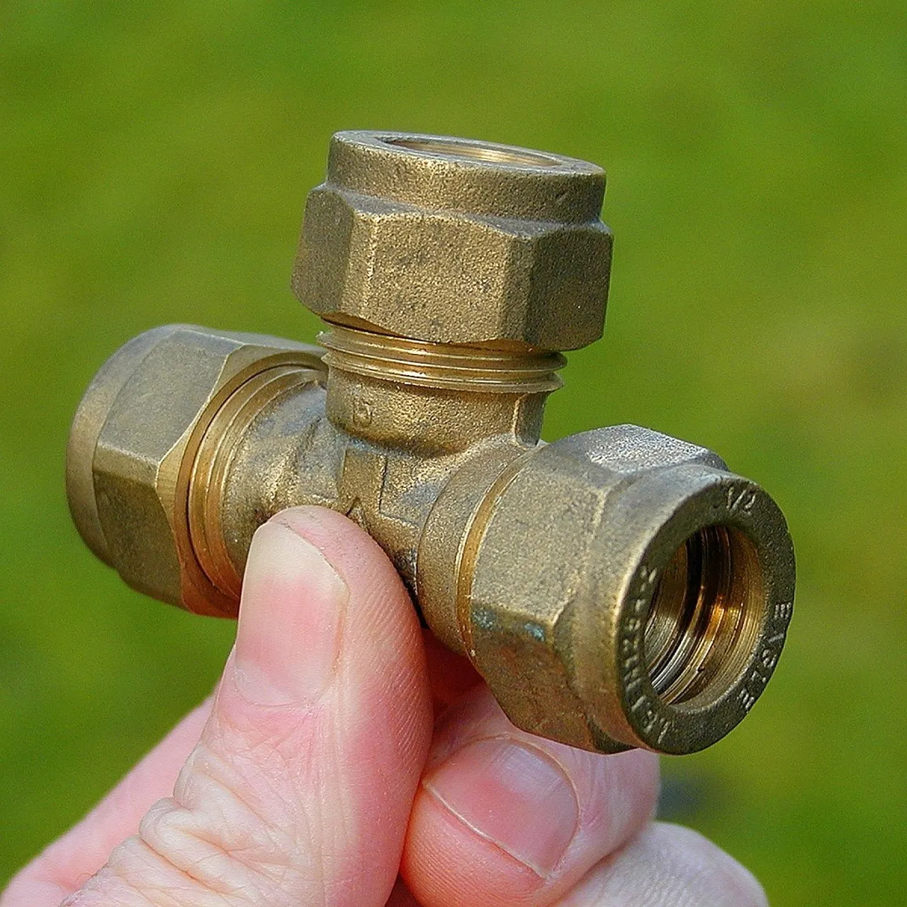 A hand holding a brass T-shaped plumbing fitting, three separate threaded ports joined into a single body