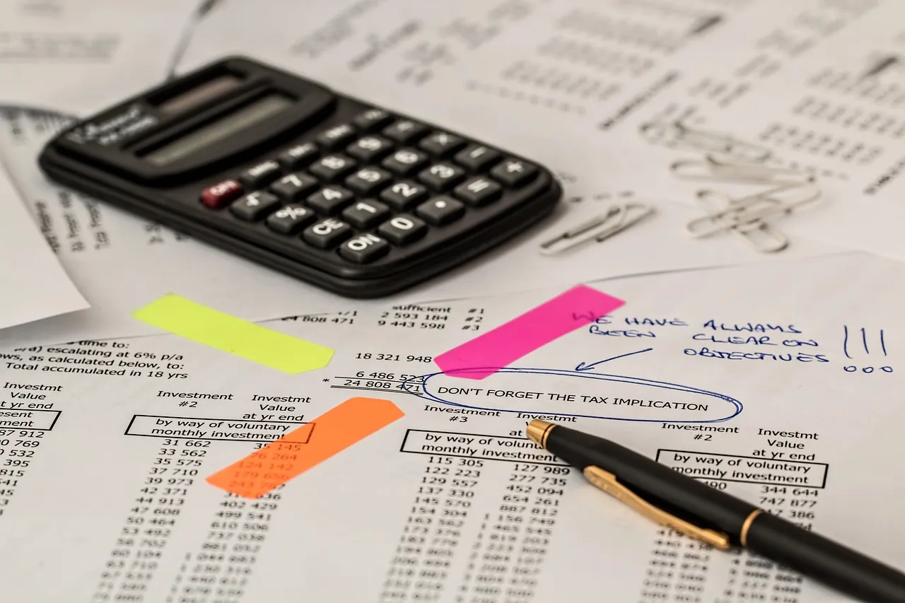 A pocket calculator and a pen resting on printed financial statements marked with pink, orange and yellow highlighter, with paper clips scattered beside them and a handwritten note circled on the page.