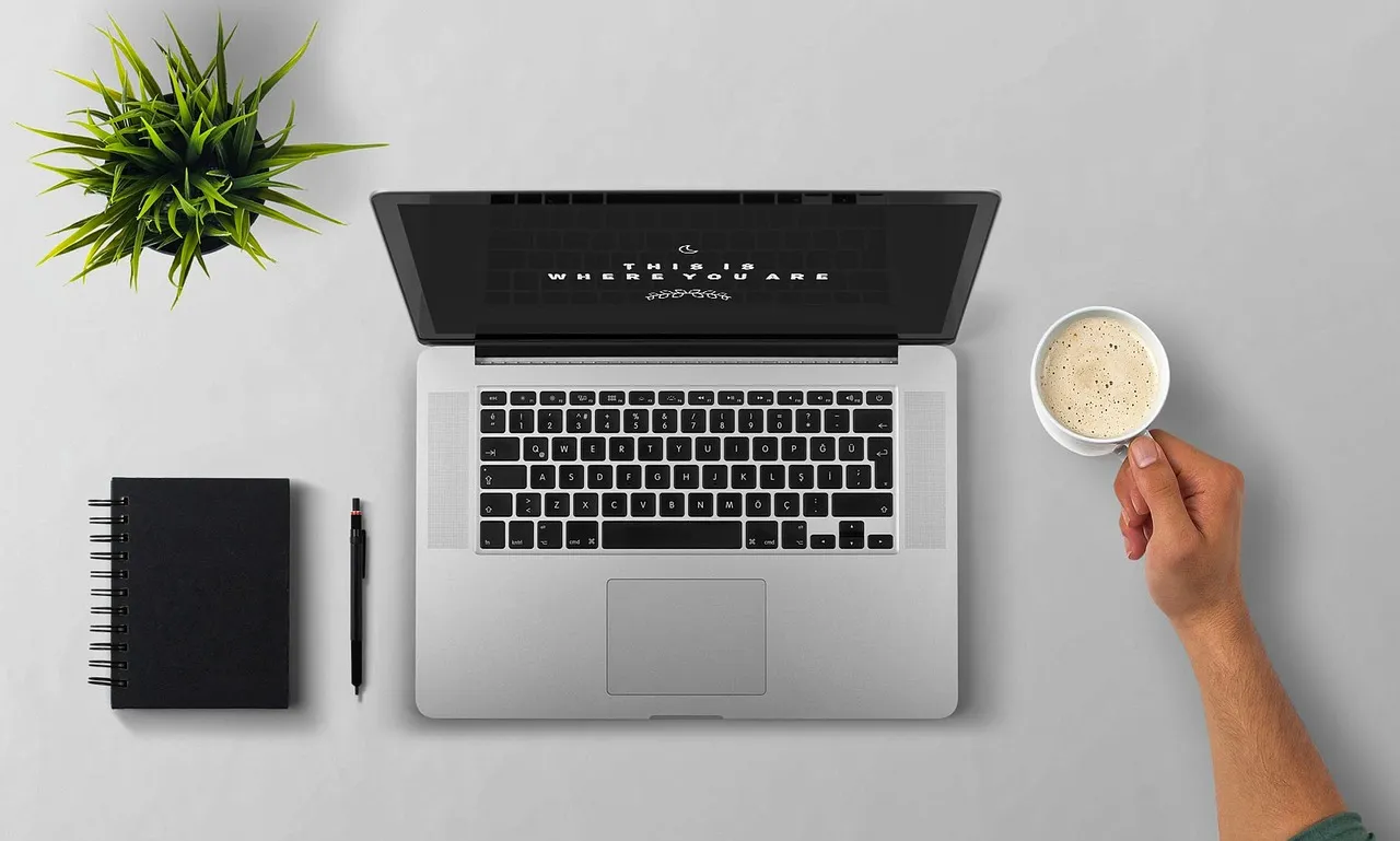 A laptop seen from above on a desk beside a notebook, pen and coffee cup