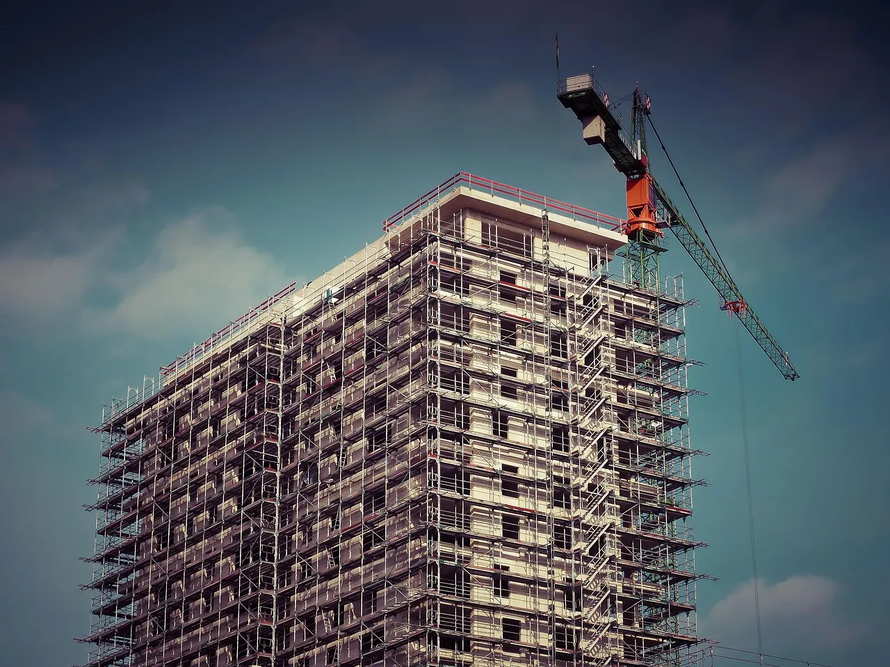 A tower block under construction, with a crane standing on the unfinished top floor against a blue sky.