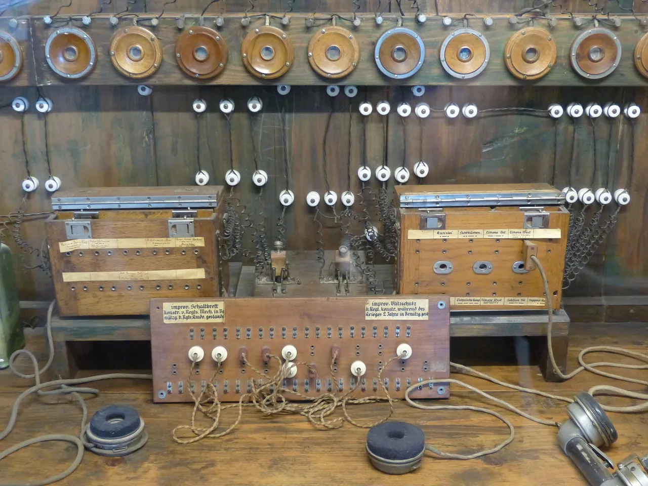 An antique wooden telephone switchboard, its rows of brass jack sockets and cloth-covered cables still wired into place, with two handset earpieces resting on the bench in front of it.