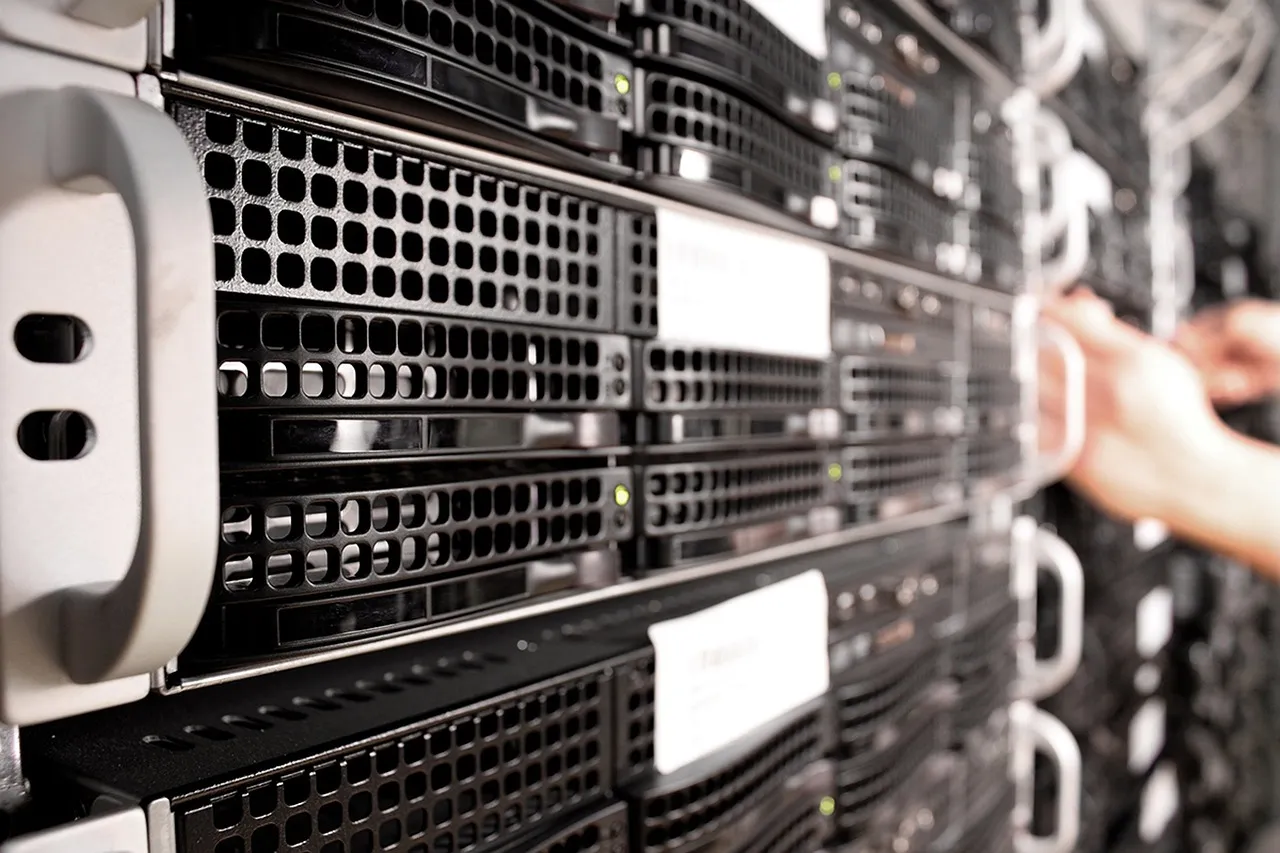 Rows of servers in a darkened data-centre rack, a hand reaching into one unit — illustrating the hosted reporting platform whose compromise reached its customers' databases.