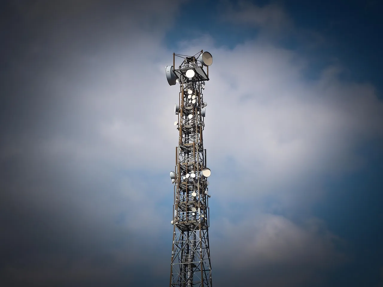 A telecommunications tower carrying panel antennas and microwave dishes against a cloudy sky