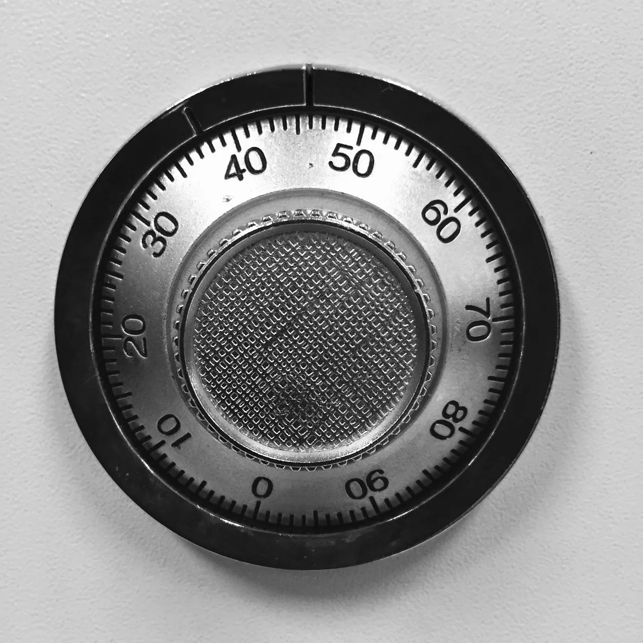 A close-up of a brushed steel combination dial set into a pale safe door, its numbered ring running from zero to ninety around a knurled centre knob.