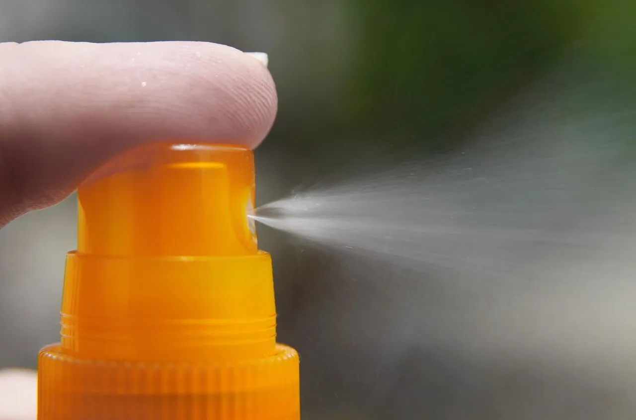 A finger pressing the nozzle of an orange sunscreen spray bottle, releasing a fine mist